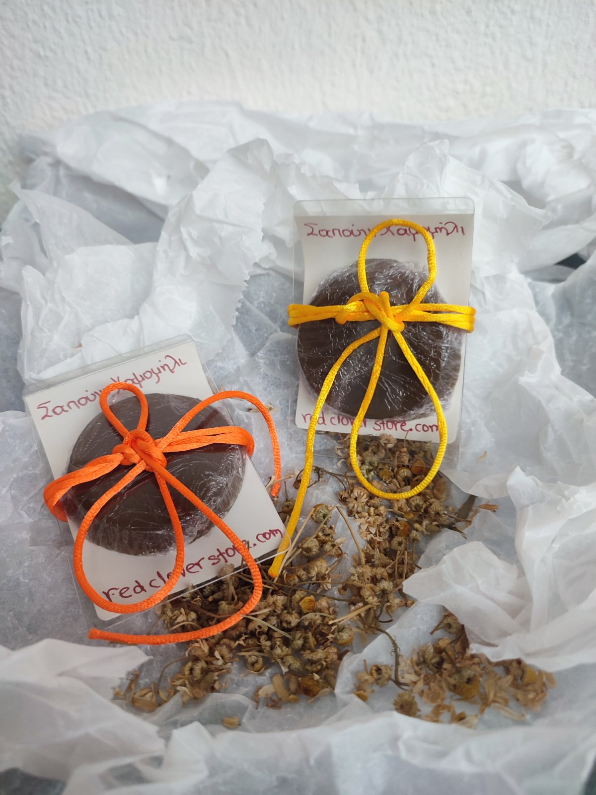 Traditional Greek chamomile and olive oil soap bars wrapped with orange and yellow ribbons, sitting on raw dried chamomile flowers.